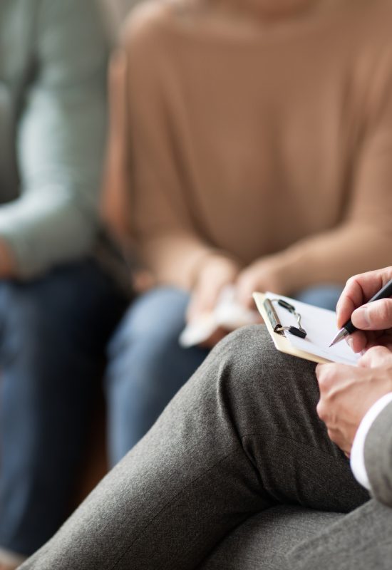 Help Concept. Closeup Cropped View Of Consultant Writing Taking Notes, Selective Focus On Hand With Pen. Spouses Having Session With Male Marriage Therapist, Sitting On Couch In Office.